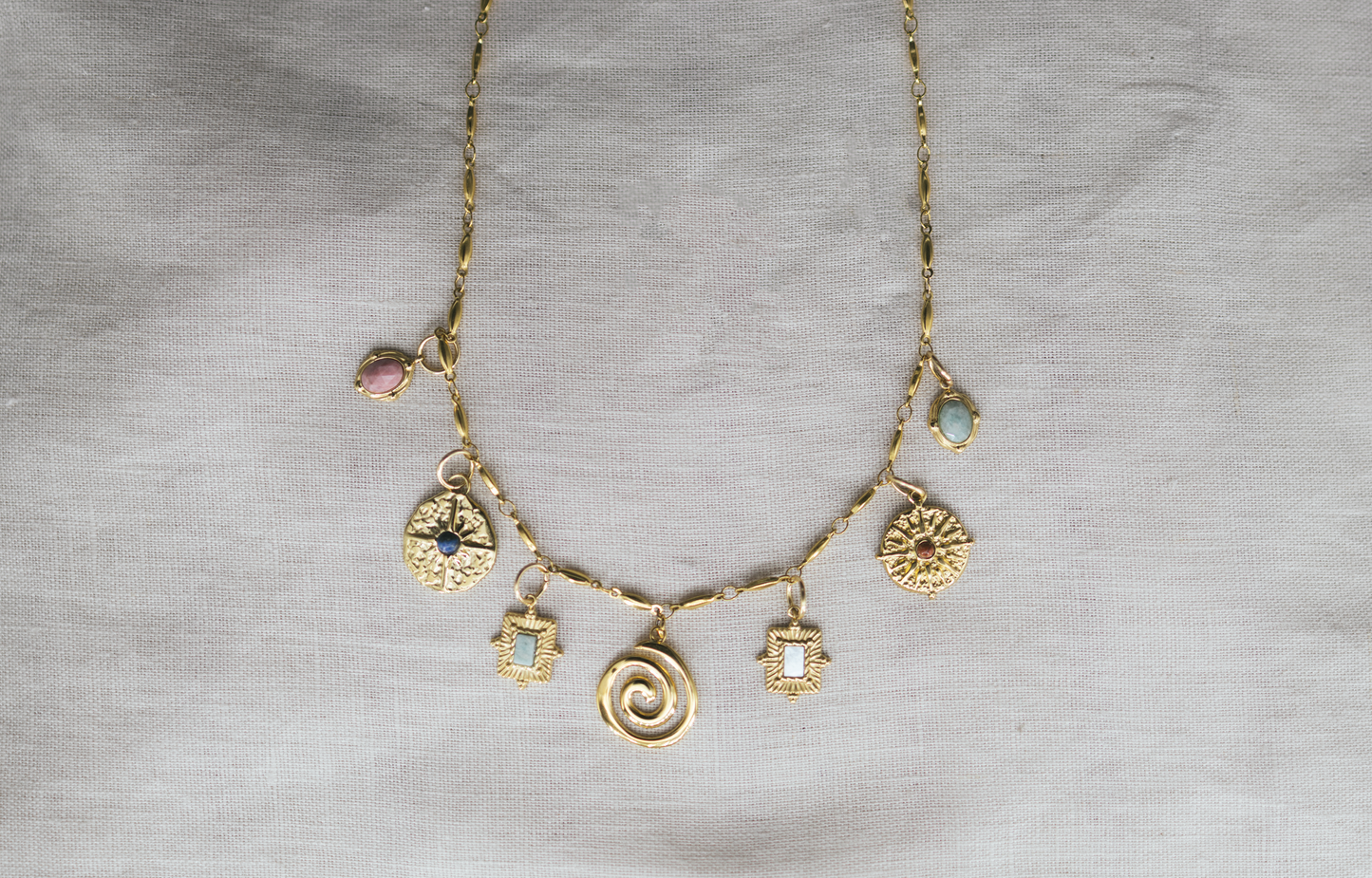 Golden Aura Charm Necklace. Gold necklace with various charms on a gray fabric background