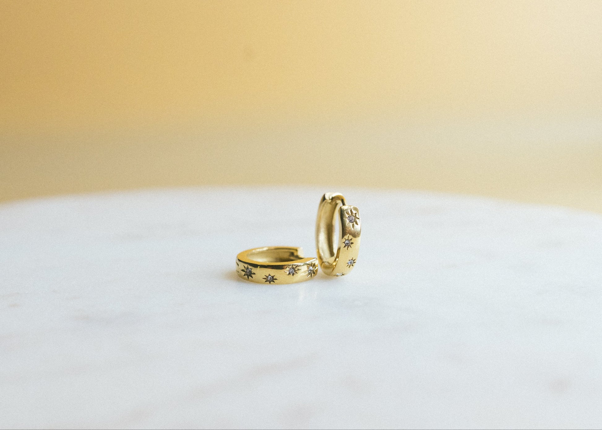 Sunbeam Glow Huggies. Two gold rings on a white surface with a beige background