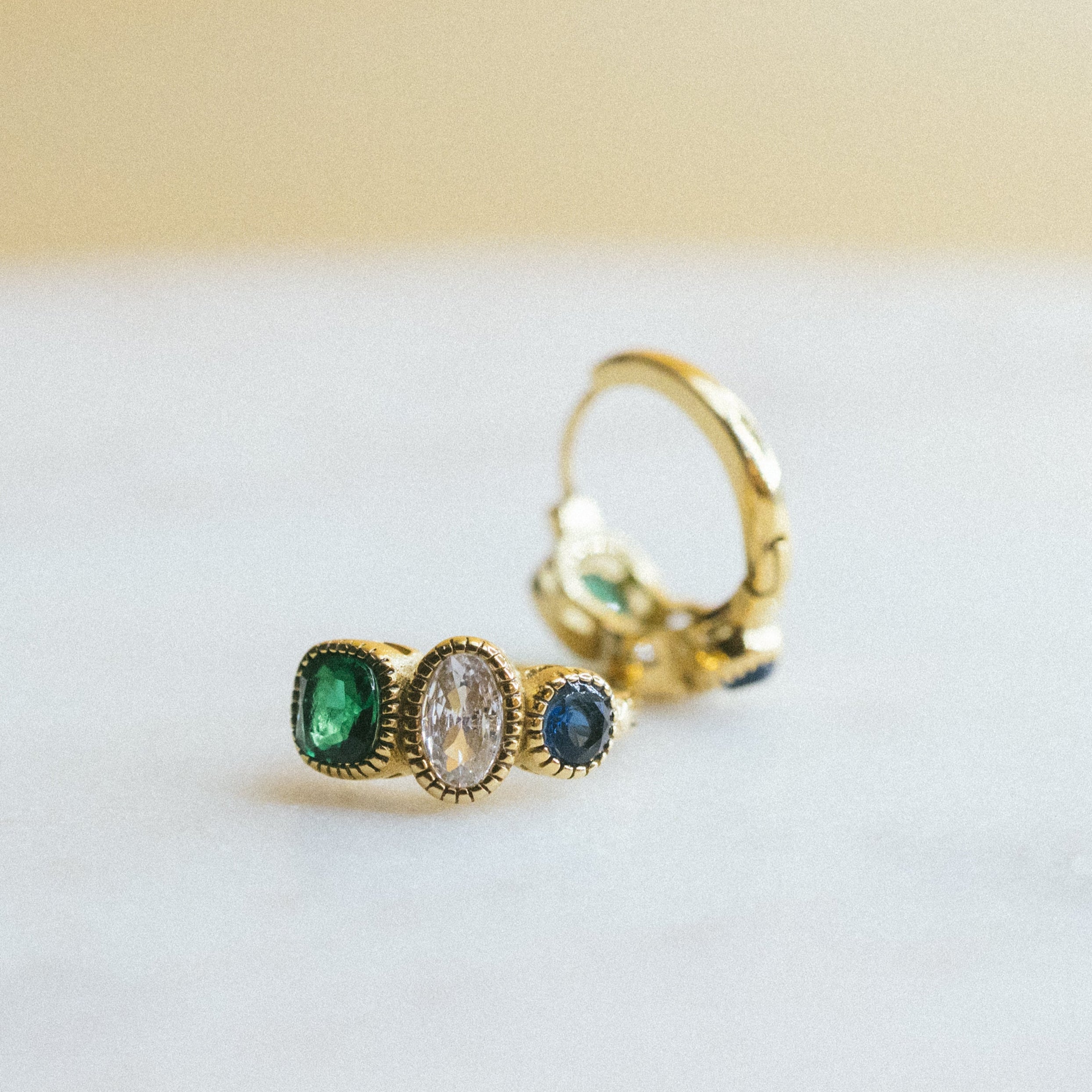 gold huggies earrings. Gold ring with green and blue gemstones on a light background